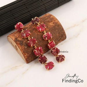 May include: A pair of long drop earrings featuring multiple raw, deep red ruby-like stones set in gold-toned metal prongs. Each earring consists of a vertical chain of five irregular, textured stones, creating a dangling, linear design.