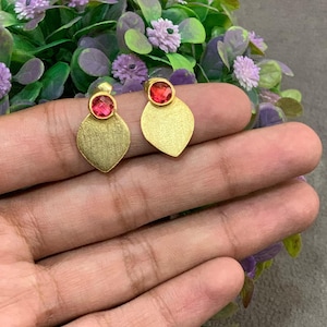 May include: A pair of gold-coloured stud earrings. Each earring has a round, red gemstone above a leaf-shaped, textured gold base. The earrings are shown against a backdrop of purple and green foliage.