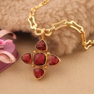 Puede incluir: Un collar dorado con un colgante en forma de cruz. El colgante presenta cinco piedras preciosas facetadas de color rojo intenso engastadas en un marco dorado. La cadena también es dorada y tiene un diseño de eslabones único. El collar se muestra sobre una superficie texturizada de color marrón claro.