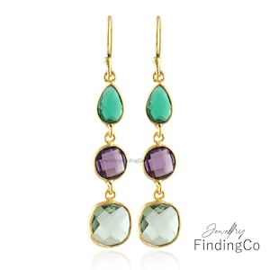 May include: Gold-toned dangle earrings with three colorful gemstones. Each earring features a green teardrop-shaped stone, a purple round stone, and a light green square stone. The earrings are from Jewellery Finding Co.