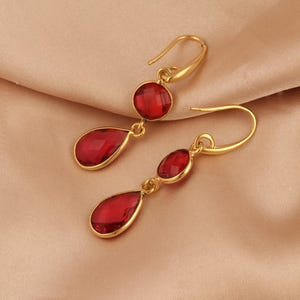 May include: A pair of gold-coloured drop earrings with red, faceted, teardrop-shaped and circular gemstones. The earrings have a hook closure and are displayed on a neutral-coloured fabric.