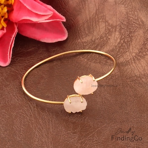 Könnte beinhalten: Goldenes Armreif-Armband mit zwei rosafarbenen Rosenquarzsteinen. Das Armband ist offen und hat ein schlichtes Design. "JewelleryfindingCo" ist auf dem Armband aufgedruckt.