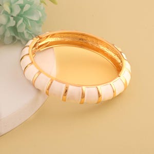 May include: A gold-tone hinged bangle bracelet with a white enamel finish and gold accents. The bracelet features a ribbed design and a secure clasp. This jewelry piece is suitable for everyday wear or special occasions.