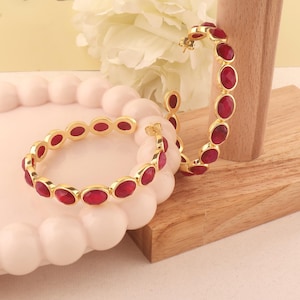 Op de afbeelding: Goudkleurige oorringen en een bijpassende armband, elk versierd met ovale, dieprode edelstenen. De sieraden worden getoond op een lichtroze, getextureerd oppervlak en een houten blok, met een witte bloem op de achtergrond.