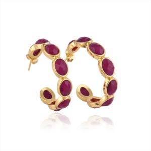 Op de afbeelding: Een paar goudkleurige oorringen, elk versierd met ovaalvormige, dieprode edelstenen. De oorbellen staan tegen een witte achtergrond, wat het contrast tussen het goud en de edelstenen benadrukt.