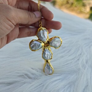 May include: A cross-shaped pendant featuring four iridescent, irregular-shaped baroque pearls set in a bright gold-toned metal frame. The pendant is suspended from a thin gold-toned chain, showcasing the natural luster and unique texture of the pearls.