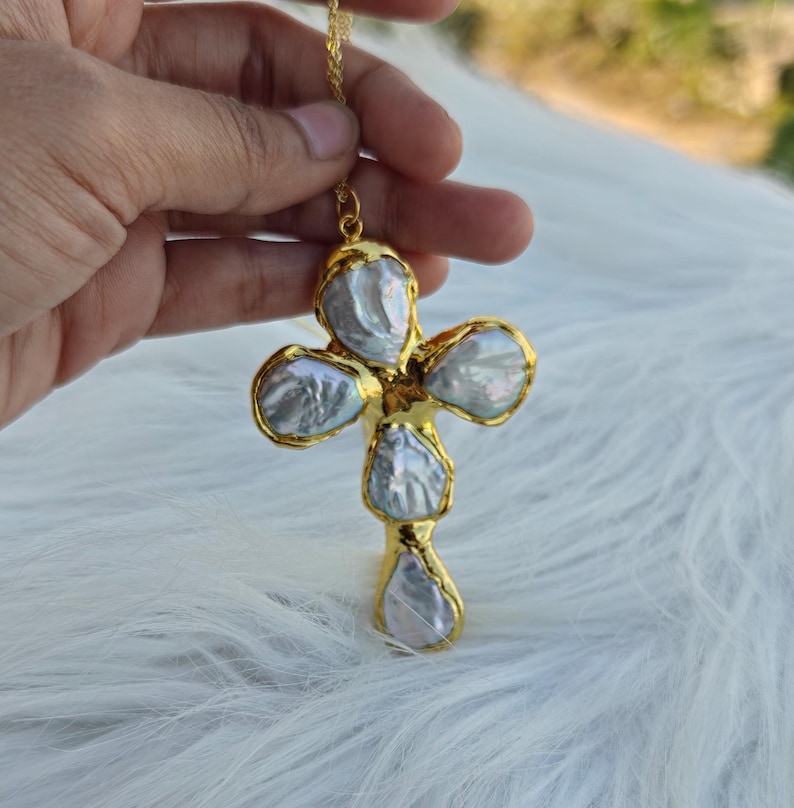 May include: A cross-shaped pendant featuring four iridescent, irregular-shaped baroque pearls set in a bright gold-toned metal frame. The pendant is suspended from a thin gold-toned chain, showcasing the natural luster and unique texture of the pearls.