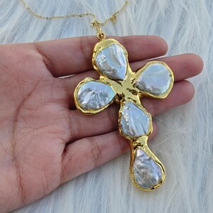 May include: A gold-toned cross pendant necklace featuring five irregular, iridescent white baroque pearls set within a textured gold metal frame. The pendant hangs from a delicate gold-toned chain, showcasing a lustrous finish on each pearl.