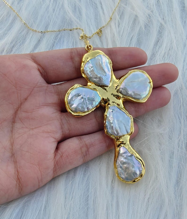 May include: A gold-toned cross pendant necklace featuring five irregular, iridescent white baroque pearls set within a textured gold metal frame. The pendant hangs from a delicate gold-toned chain, showcasing a lustrous finish on each pearl.