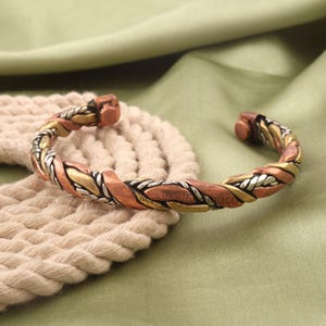 May include: A braided cuff bracelet with a mix of copper, gold, and silver tones. The bracelet is open-ended and rests on a cream-colored, woven surface against a green background. The metal bracelet has a textured, woven design.