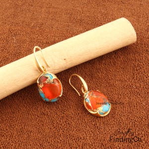 May include: Gold-coloured earrings featuring oval orange and turquoise stones. The earrings have a hook fastening and are set against a brown background with a wooden dowel.