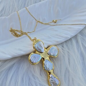 May include: A gold-toned necklace featuring a cross-shaped pendant composed of five irregular, iridescent white baroque pearls set within a polished gold metal frame, suspended from a delicate gold-toned chain with a small round bead detail.