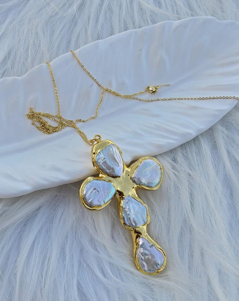 May include: A gold-toned necklace featuring a cross-shaped pendant composed of five irregular, iridescent white baroque pearls set within a polished gold metal frame, suspended from a delicate gold-toned chain with a small round bead detail.