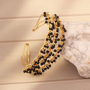 Puede incluir: Un pendiente de oreja dorado con un diseño curvo, adornado con hebras de pequeñas cuentas negras. El pendiente tiene una apariencia delicada y elegante. Esta pieza es adecuada para ocasiones especiales o para añadir un toque de glamour a un atuendo.
