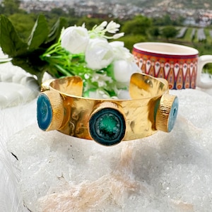 May include: A gold-colored cuff bracelet with three circular, teal-colored stone accents. The bracelet has a hammered texture and is displayed on a white surface with flowers and a decorative mug in the background. The bracelet is a piece of jewelry.