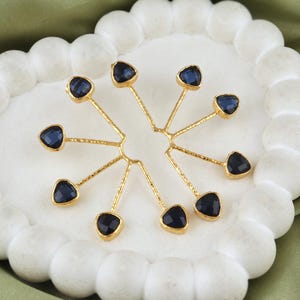 Op de afbeelding: Een cluster van goudkleurige sieraden, elk met een donkerblauwe, driehoekige edelsteen. De stukken zijn gerangschikt in een radiaal patroon, dat lijkt op een gestileerde bloem of ster. De edelstenen zijn gezet in goudkleurig metaal.