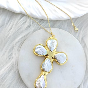 May include: A gold-toned necklace featuring a cross-shaped pendant composed of five irregular, lustrous white baroque pearls set within a polished gold metal frame. The pendant hangs from a delicate gold-toned chain with a lobster claw clasp.