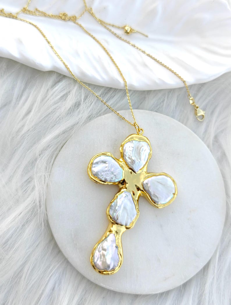 May include: A gold-toned necklace featuring a cross-shaped pendant composed of five irregular, lustrous white baroque pearls set within a polished gold metal frame. The pendant hangs from a delicate gold-toned chain with a lobster claw clasp.