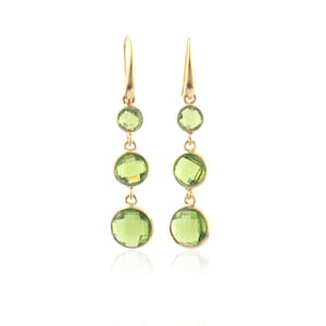 Puede incluir: Un par de pendientes colgantes dorados con tres piedras preciosas redondas de color verde claro de diferentes tamaños. Los pendientes tienen un diseño sencillo con cierre de gancho.