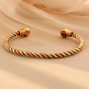 Könnte beinhalten: Ein verdrehtes Metall-Armreif-Armband in drei Farben. Das Armband hat ein geflochtenes Muster aus gold-, silber- und kupferfarbenen Metallsträngen. Die Enden des Armbands haben eine goldfarbene Kappe mit kupferfarbenem Zentrum.