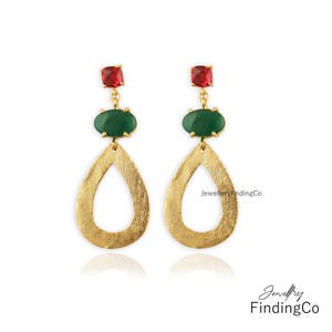 May include: A pair of gold-coloured drop earrings. Each earring features a red square gemstone at the top, a green oval gemstone in the middle, and a textured, teardrop-shaped gold pendant. The earrings are displayed on a white background.