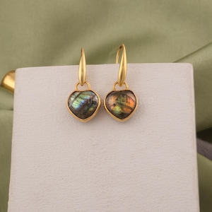 May include: A pair of gold-toned drop earrings featuring heart-shaped labradorite gemstones. Each earring has a smooth gold metal hook and a bezel setting that secures the iridescent, dark-toned stone, displaying flashes of green and blue light.