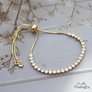 Puede incluir: Una pulsera ajustable dorada con una fila de gemas claras y brillantes. La pulsera tiene una cadena delicada y una cuenta deslizante para ajustar. El texto "Jewellery FindingCo" es visible en la imagen.