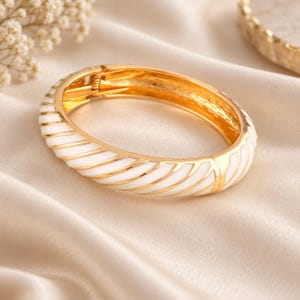 May include: A hinged bangle bracelet featuring a polished gold-tone metal frame with diagonal white enamel stripes. The bracelet has a secure clasp mechanism and a smooth, rounded profile, showcasing a repeating pattern of gold and white segments.