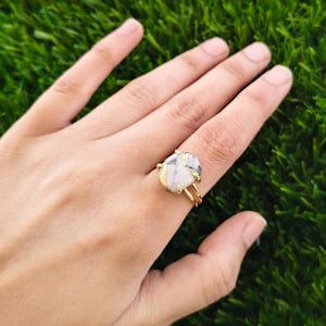 Pode incluir: Um anel de tom dourado com uma pedra central irregular branca com veios cinzentos. A pedra é mantida por uma cravação de metal dourado que envolve as bordas, ligada a um aro fino de metal dourado com banda dupla.