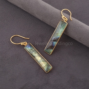 Könnte beinhalten: Ein Paar goldfarbene, rechteckige Ohrringe mit schillernden, blau-grünen Steinen. Die Ohrringe haben ein Haken-Design für durchbohrte Ohren. Die Steine sind in einen goldfarbenen Metallrahmen gefasst. Die Ohrringe werden auf einer grauen Oberfläche präsentiert.