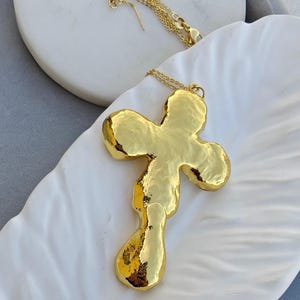 May include: A gold-toned necklace featuring a large, irregular, organic-shaped cross pendant with a high-polish, hammered-like texture. The pendant is suspended from a delicate, thin gold-toned chain with a small lobster claw clasp closure.
