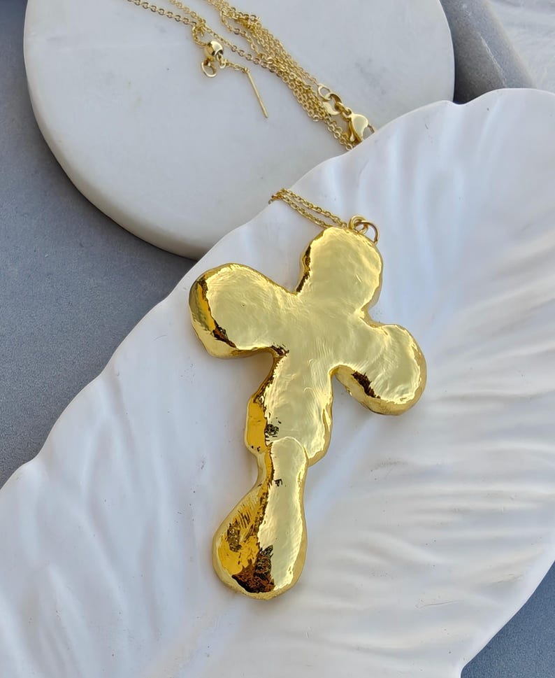 May include: A gold-toned necklace featuring a large, irregular, organic-shaped cross pendant with a high-polish, hammered-like texture. The pendant is suspended from a delicate, thin gold-toned chain with a small lobster claw clasp closure.