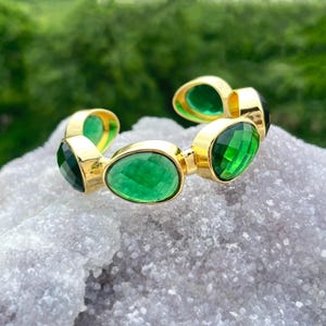Könnte beinhalten: Ein goldfarbenes Armband mit mehreren ovalen und smaragdgeschliffenen grünen Edelsteinen. Die Edelsteine sind in goldenen Fassungen gefasst, was einen auffälligen Kontrast erzeugt. Das Armband wird auf einer hellgrauen, strukturierten Oberfläche mit verschwommenem grünem Hintergrund präsentiert.