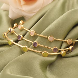 May include: Three gold bangles with colourful gemstones. Each bangle features a series of round gemstones in shades of pink, purple, and yellow-green. The bangles are displayed on a green satin fabric.