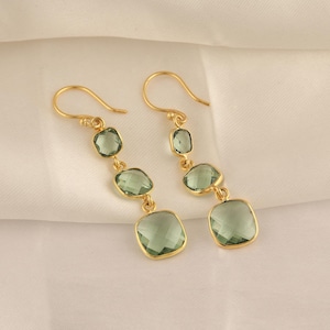 May include: Gold-tone earrings with a series of three green-hued, faceted gemstones. The gemstones are in a mix of shapes, including round and square, and are set in gold bezels. The earrings hang from gold-colored ear wires.