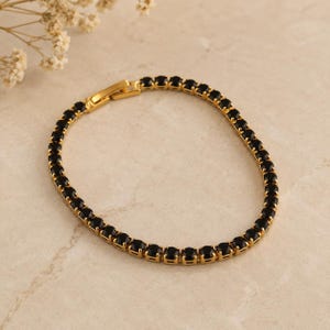 May include: A gold-toned tennis bracelet featuring a continuous row of round, faceted black stones set in a metal frame. The bracelet includes a secure, rectangular-shaped clasp mechanism that connects the two ends of the circular jewellery piece.