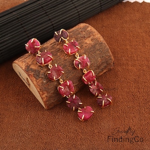 Può includere: Un paio di orecchini lunghi e pendenti con più perline di pietre preziose rosse collegate da un filo d'oro. Gli orecchini sono su una superficie di legno marrone.