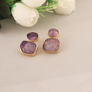 Pode incluir: Um par de brincos dourados com pedras de ametista em bruto de formato irregular. Os brincos têm um design de duas pedras, com uma pedra menor em cima de uma maior. As pedras de ametista exibem tons de roxo. Fundo neutro.