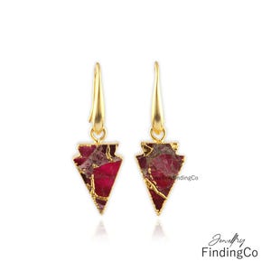 May include: A pair of gold earrings with red and gold arrow-shaped pendants. The earrings are made of gold-plated metal and feature a red and gold stone.