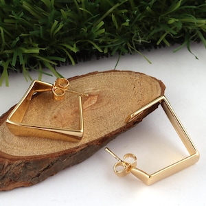 May include: A pair of gold-coloured square earrings. The earrings have a geometric design, with a post and butterfly backing. The earrings are displayed on a piece of wood with a green background.