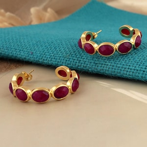 Puede incluir: Un par de pendientes de aro dorados, cada uno adornado con piedras preciosas ovaladas de color rojo intenso. Los pendientes se exhiben sobre una tela turquesa, mostrando su vibrante color y diseño. Las piedras están engastadas en metal dorado.
