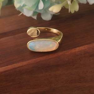 May include: A gold ring featuring an oval opal gemstone. The ring has an open design with a small, circular accent. The opal displays a play of colours, including white, blue, and yellow. The ring is a piece of jewellery.
