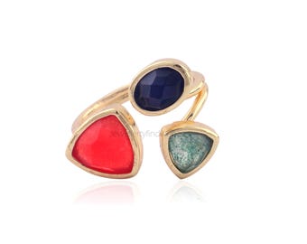 May include: Gold-toned ring featuring three colorful gemstones. The ring has a red triangular stone, a blue oval stone, and a green triangular stone. The gemstones are set in a gold-colored metal.