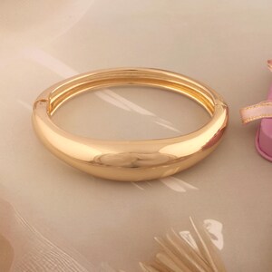 Può includere: Un bracciale a cerchio dorato, incernierato, dal design liscio e arrotondato. Il bracciale ha una finitura lucida e uno stile semplice ed elegante. Il gioiello è esposto su una superficie colorata chiara.