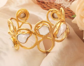 Puede incluir: Un brazalete tipo puño de tono dorado con un diseño calado de marcos metálicos circulares interconectados. Varias perlas planas, blancas y lustrosas de forma irregular están engastadas en las aberturas circulares de la estructura.
