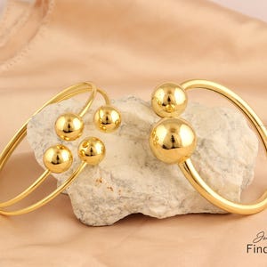 Puede incluir: Un conjunto de tres pulseras de brazalete doradas. Una pulsera presenta una sola bola grande, mientras que la otra pulsera presenta tres bolas más pequeñas. Las pulseras se muestran en una roca.