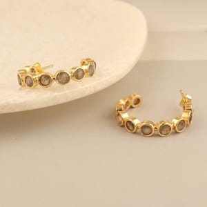 Peut inclure: Une paire de boucles d'oreilles créoles en or avec une rangée de pierres ovales en labradorite.