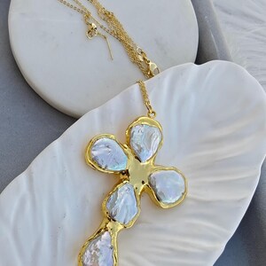 May include: A gold-toned necklace featuring a cross-shaped pendant composed of five irregular, iridescent white baroque pearls set within a polished gold metal frame. The delicate gold chain is attached to the top of the cross pendant.