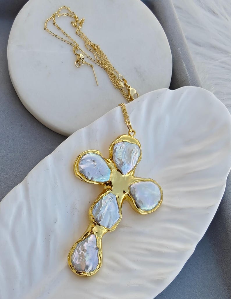 May include: A gold-toned necklace featuring a cross-shaped pendant composed of five irregular, iridescent white baroque pearls set within a polished gold metal frame. The delicate gold chain is attached to the top of the cross pendant.