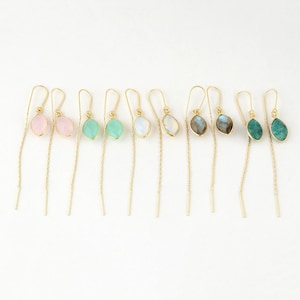 May include: A collection of threader earrings with gold-colored chains and various gemstone pendants. The earrings feature pink, green, white, and blue-green teardrop-shaped stones. The earrings are displayed on a white background.